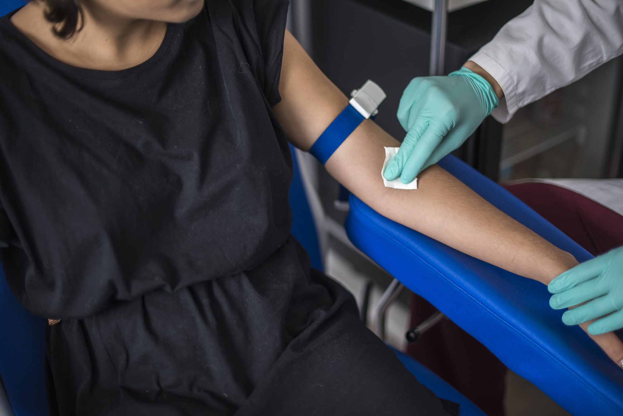 Braccio con laccio emostatico blu disinfettato con garza da mani in guanti verdi, appoggiato su un bracciolo blu
