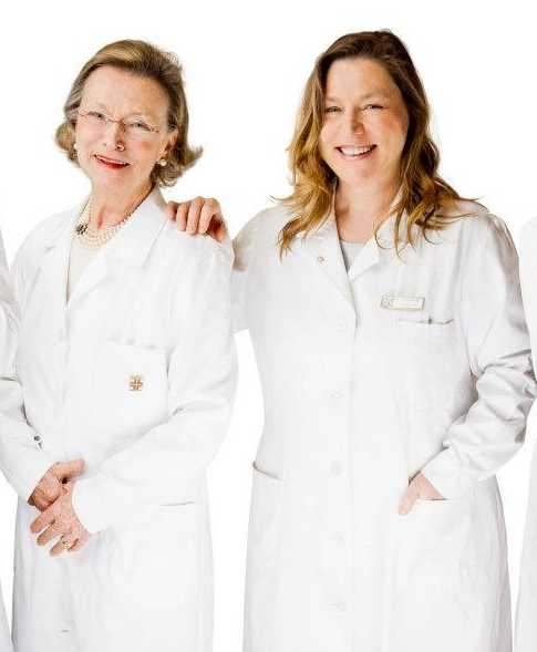 Due donne sorridenti in camice bianco su sfondo bianco, una più anziana con occhiali e collana di perle, l'altra bionda